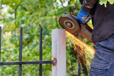 A worker cuts a metal post with a disc angle grinder, creating a bright plume of many hot sparks.