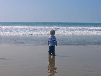 Rear view of boy standing on sea shore at beach against clear sky