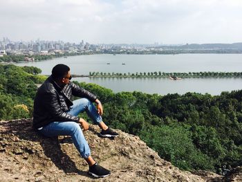 Full length of man sitting on rock against lake in city