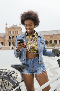 Woman using mobile phone while standing with bicycle on footpath