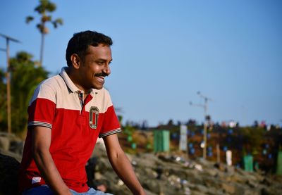 Happy man looking away while sitting by hill against clear blue sky