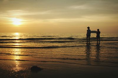 Silhouette men standing on shore at beach against sky during sunset