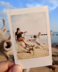 Cropped hand of woman holding instant print transfer at beach against sky