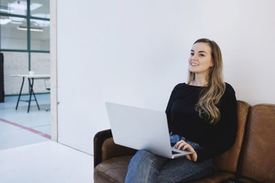 Smiling young woman looking away while sitting with laptop on sofa at office