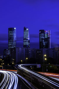 Light trails on road by illuminated buildings against sky at night