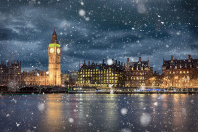 Illuminated city by river against sky at night during snowfall