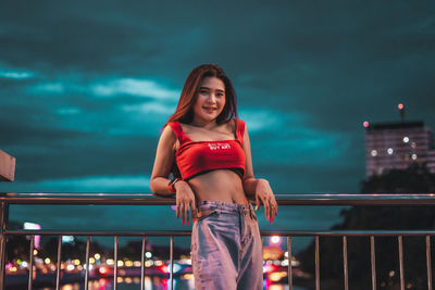 Low angle view of smiling young woman standing on bridge against sky at night