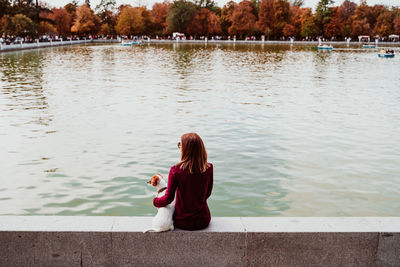 Rear view of woman with dog sitting by lake against trees