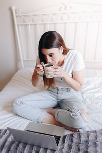 Young woman drinking coffee while using laptop on bed at home