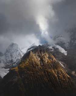 Dramatic view of forested high rocky slopes and snowy mountains in gray cloudy sky in changeable