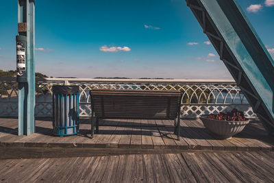 Empty bench by garbage bin on floorboard against sky