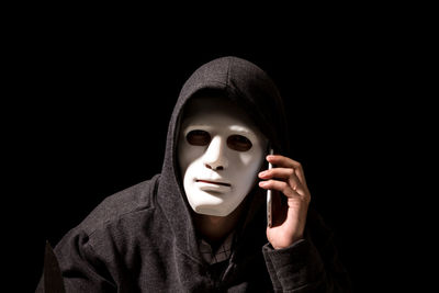 Portrait of criminal in mask using mobile phone against black background