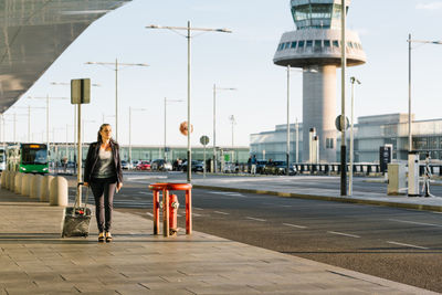 Cheerful female tourist with suitcase and backpack walking in airport and smiling away while enjoying summer weekend