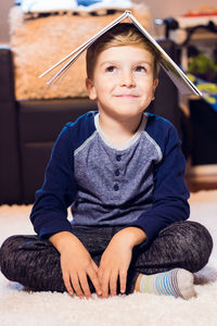 Thoughtful boy with book on head sitting at home