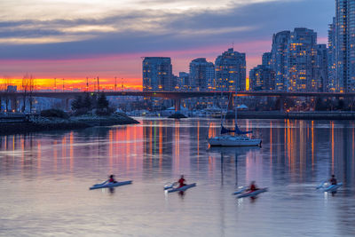 Boats in river by buildings against sky during sunset