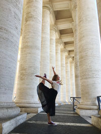 Side view of woman with arms raised standing on footpath against columns