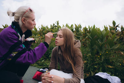 Young woman doing make-up on friend sitting on grass against sky