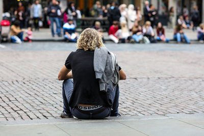 Rear view of young man sitting on footpath on crowded street