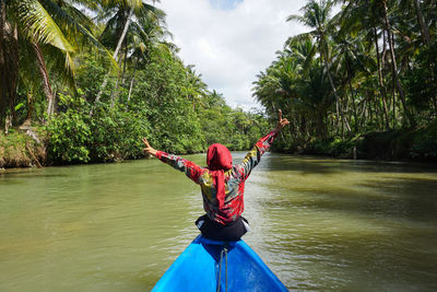 Rear view of woman with arms outstretched sitting on boat over river against sky