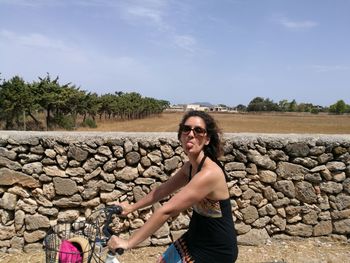 Portrait of woman sticking out tongue while riding bicycle by stone wall