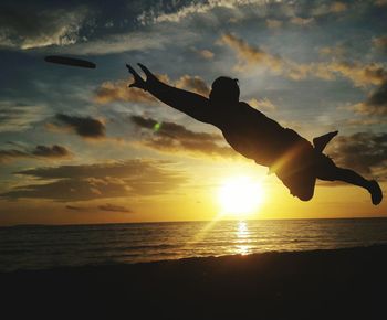 Silhouette man catching plastic disc at beach against sky during sunset