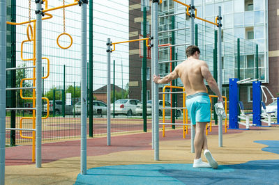 Rear view of shirtless man standing by gymnastics equipment against building