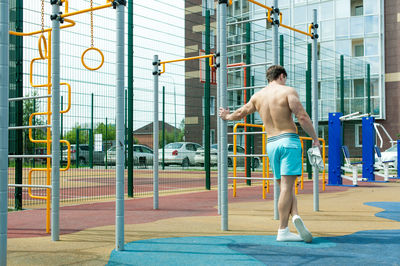 Rear view of shirtless man standing by gymnastics equipment against building