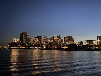 Illuminated buildings by river against clear sky at dusk