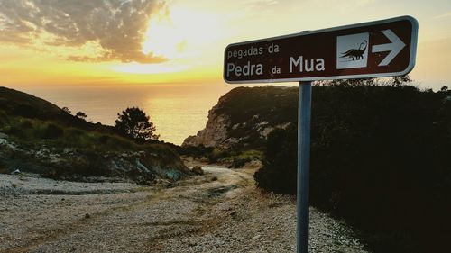 Road sign by sea against sky during sunset