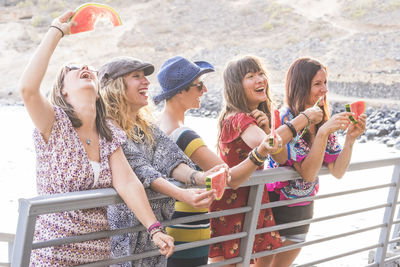 Happy friends eating watermelon while standing by railing