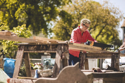 Senior woman cutting wooden plank with hand saw in yard