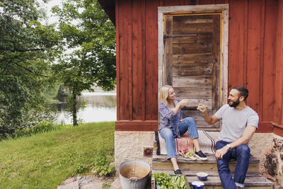 Happy friends toasting drink while sitting outside cottage against lake