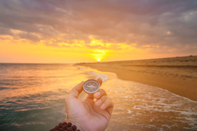 Cropped hand holding navigational compass at beach against sky during sunset