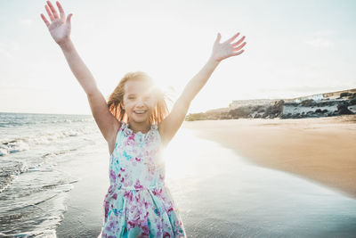 Portrait of smiling girl with arms raised standing at beach against sky