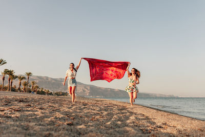 Cheerful female friends holding scarf and running at beach