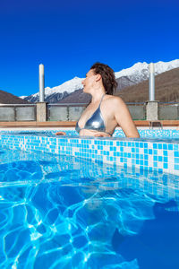 Slender woman in a silver bikini in winter by outdoor pool against the mountains and clear blue sky