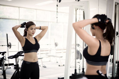 Young woman tying hair looking in mirror while standing at gym