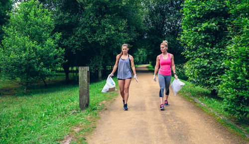 Full length of friends walking on footpath while holding plastic bags against trees