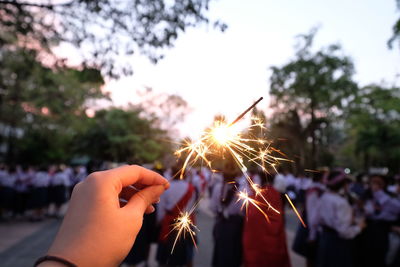 Cropped hand holding lit sparkler with crowd on road in city during sunset