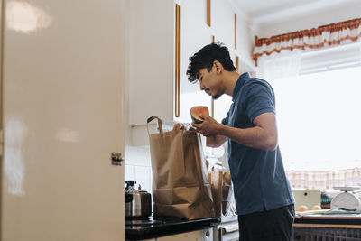 Side view of male care assistant unpacking groceries from bag in kitchen at home