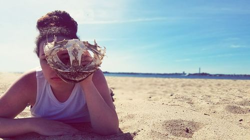 Portrait of young woman holding animal skull while lying at beach against sky during sunny day