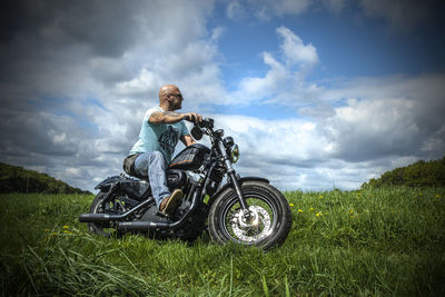 Low angle view of man with motorcycle on grassy field against cloudy sky