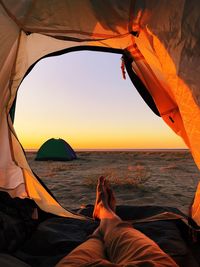 Low section of person lying in tent at beach against sky during sunset