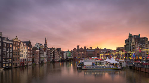 Boats in river by buildings against sky during sunset