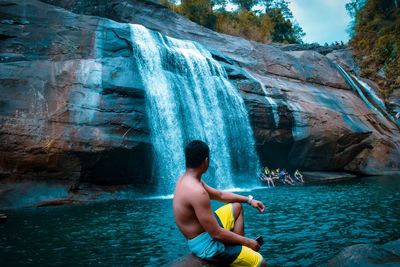 Shirtless man sitting against waterfall