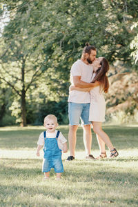 Baby boy standing on grass land while parents kissing in background at park