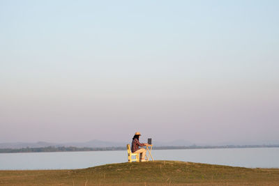 Woman using laptop over table sitting on grass by lake against clear sky