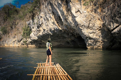 Side view of woman standing on wooden raft in sea against rock formation