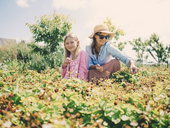 Smiling woman and daughter sitting amidst plants at farm against sky on sunny day