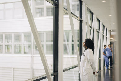 Side view of female physician looking through window while standing in corridor at hospital
